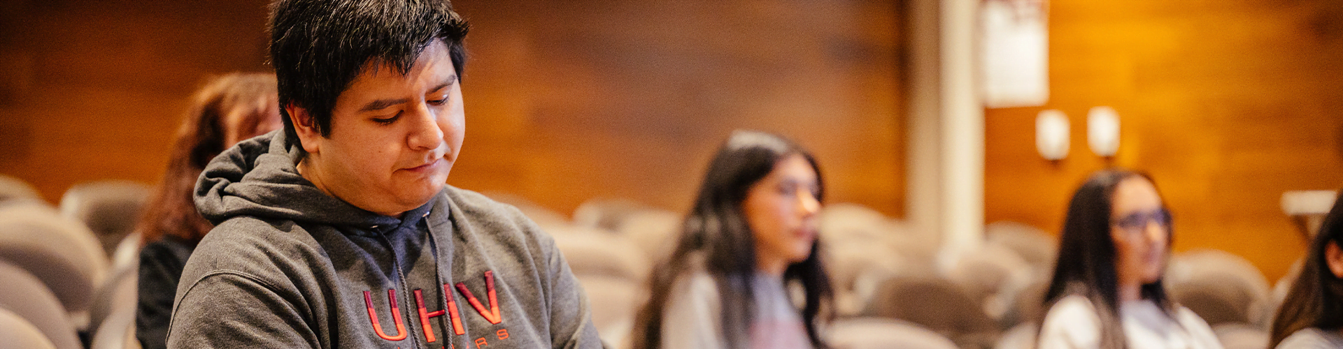 People sitting in an auditorium or classroom, focus on person in grey hoodie with red text 'UHV'
