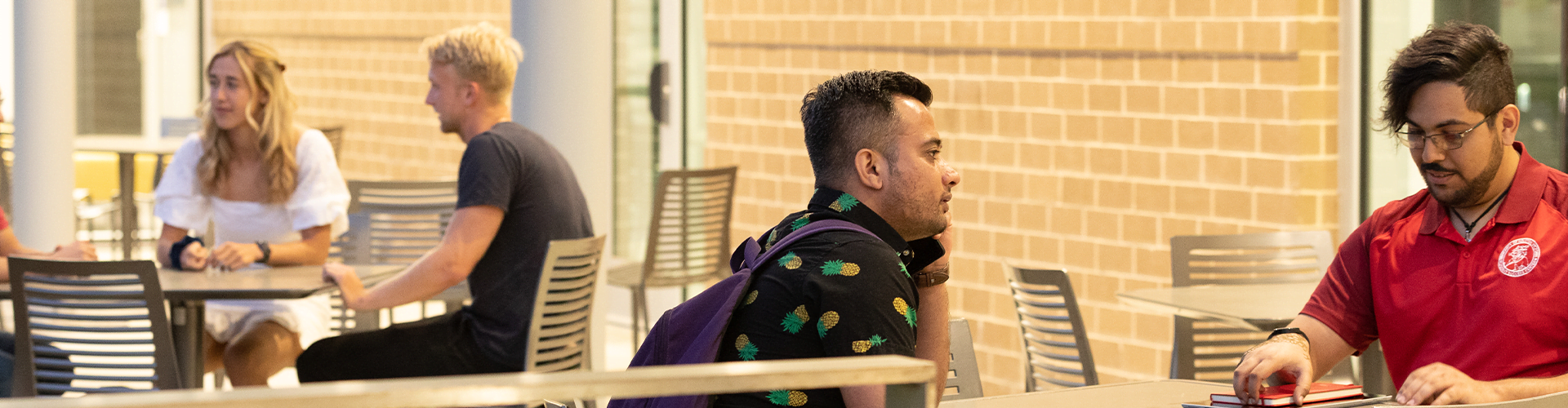 Three people sitting at tables indoors—one in a black shirt with green patterns and a purple backpack, one in a red shirt, and one in a white dress