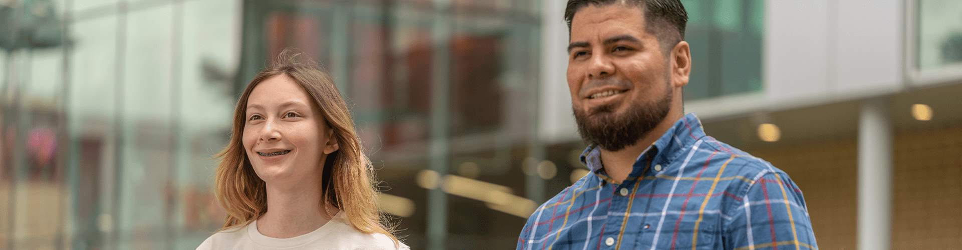 Two people standing outdoors in front of a modern building with glass windows and brickwork, one in a light top and the other in a blue plaid shirt.