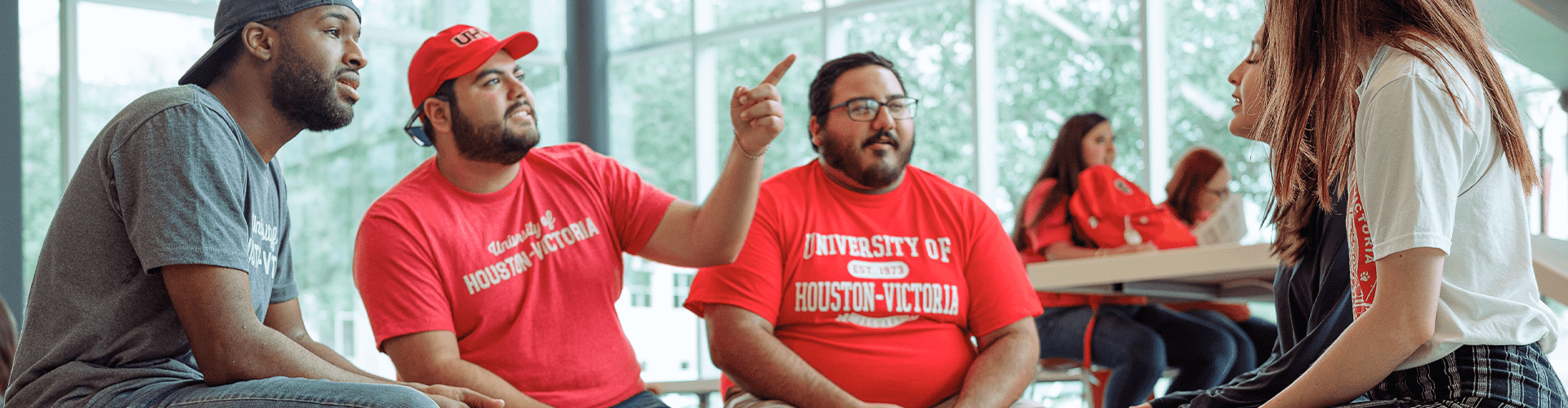 A group of people sitting and talking indoors. Two individuals in the foreground are wearing red shirts with 'UNIVERSITY OF HOUSTON-VICTORIA' printed on them.