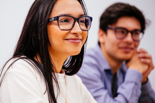 Two students sitting side by side, one wearing a white sweater and glasses, the other in a blue shirt and glasses.