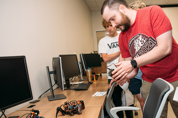Two students collaborating on a robotics project in a computer lab, with one examining a small robot on a desk surrounded by computer monitors