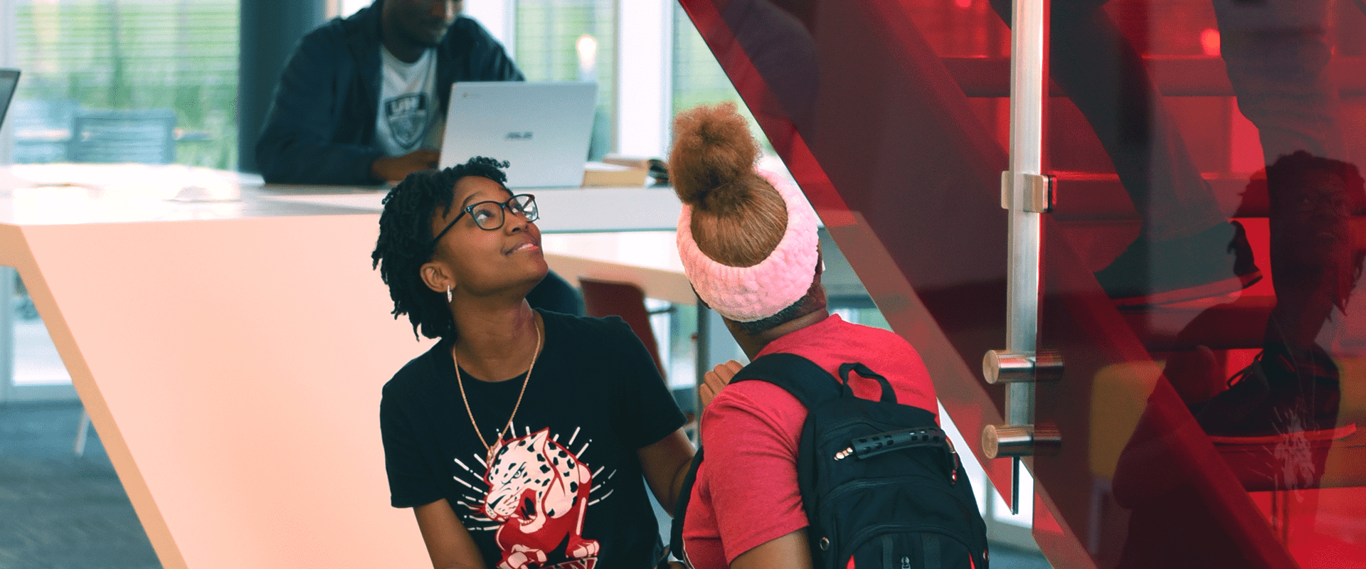 Two students in the UHV South building, watching others walk up the stairs lined with red glass walls.