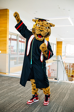 Jax, the University of Houston-Victoria jaguar mascot, wearing a graduation gown and cap, with one arm raised in celebration.