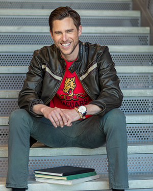 Person in UHV Jaguars shirt and leather jacket sitting on stairs with books