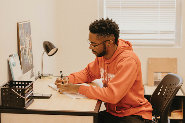 Student in an orange hoodie writing at a desk in a dorm room with study materials and a lamp