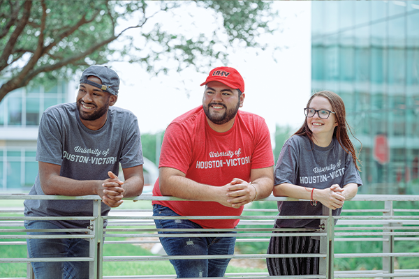 Three people leaning on a metal railing outdoors, wearing UHV t-shirts. Trees and buildings are visible in the background.