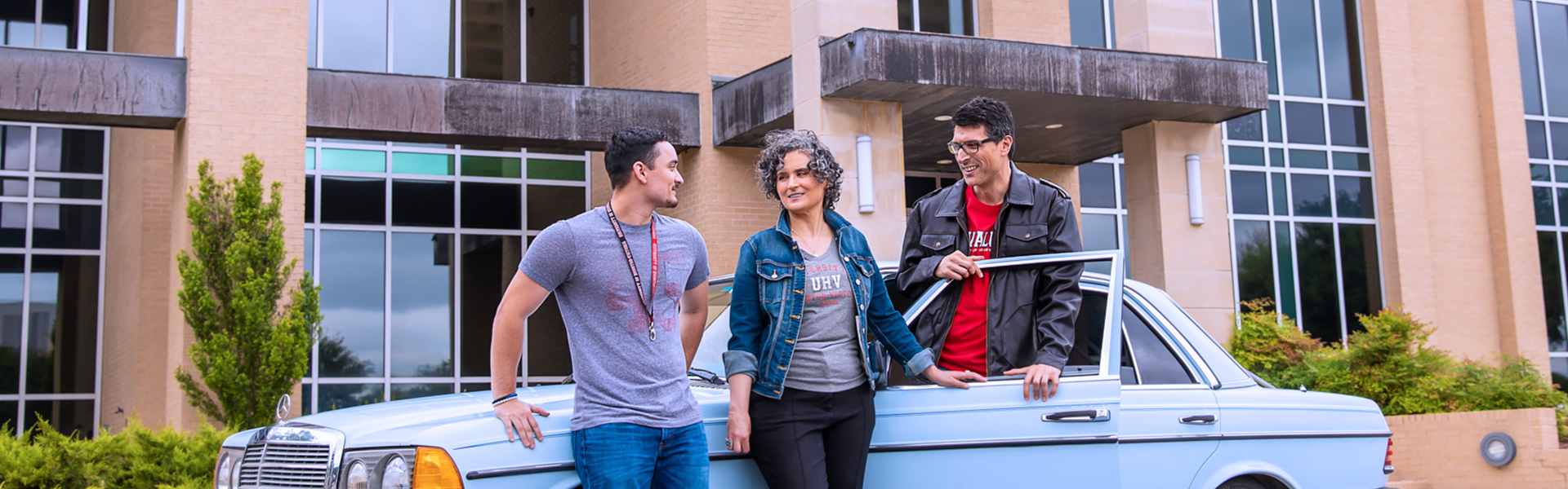 A group of people standing in front of the University West building, leaning against a classic light blue Mercedes-Benz from the 1970s or 1980s. They are dressed casually in jeans and jackets.