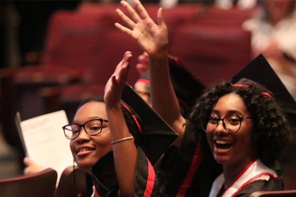 Two students at a graduation ceremony, smiling and waving to onlookers.