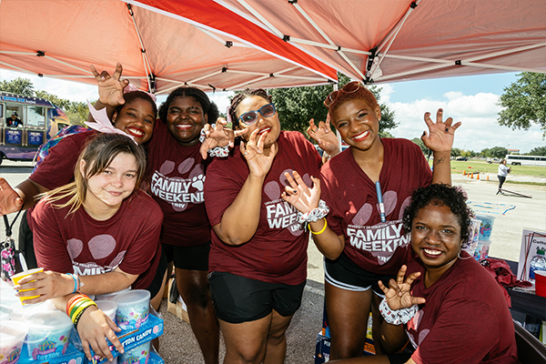 A group of students under a red canopy tent wearing matching “UHV Family Weekend” shirts, showing the jaguar hand-sign