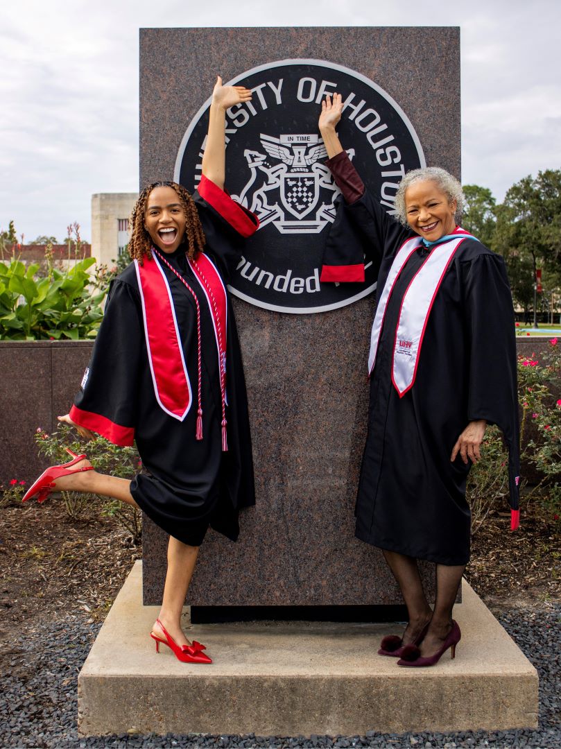 Two women in graduation robes stand in front of the University of Houston seal.