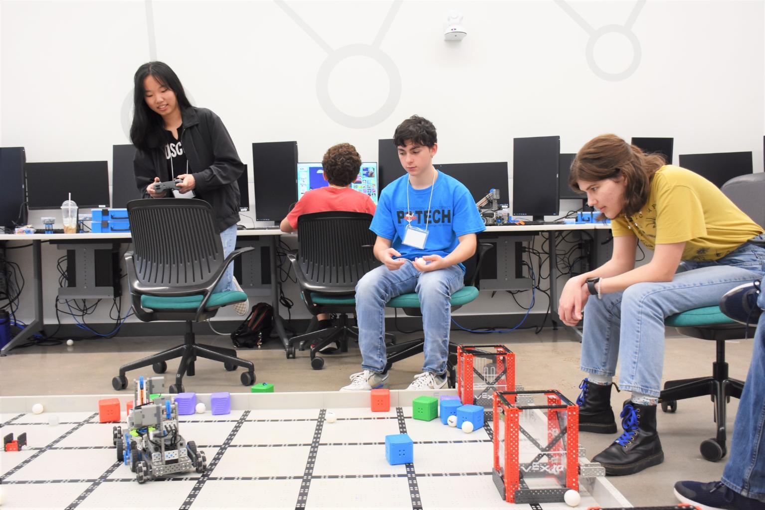 A high school student controls a small robot while two other students sit and watch.