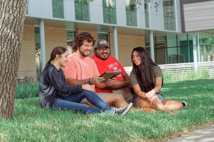A group of four UHV students siting outside UHV South building looking at a tablet