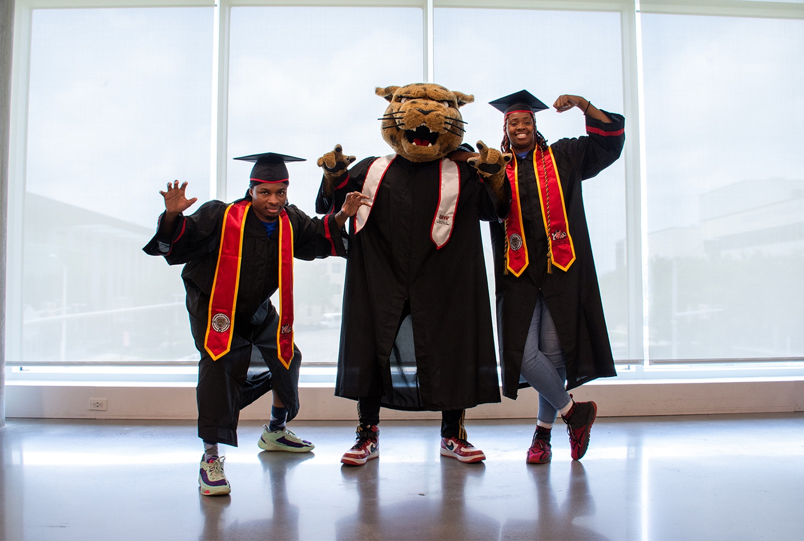 jaX the jaguar mascot with two graduates in black gowns and red and gold stoles.